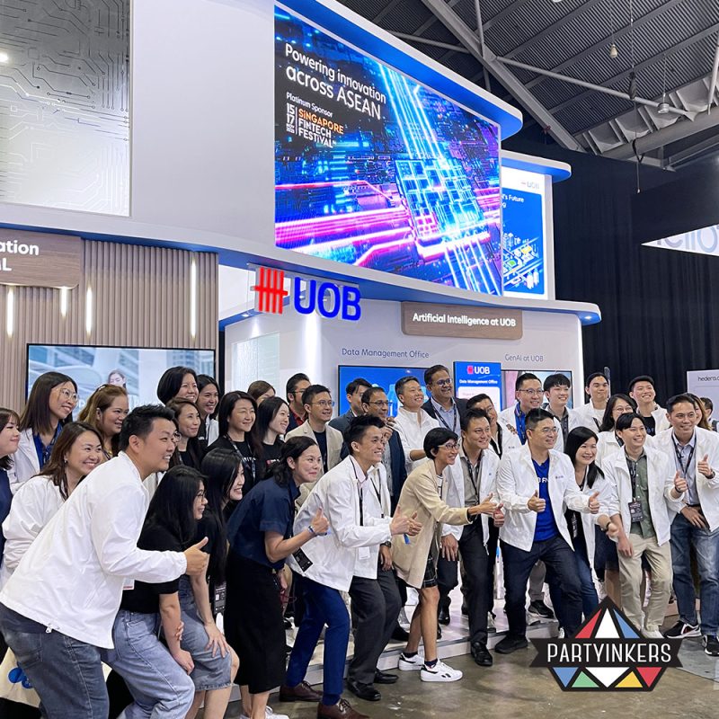 Group of individuals posing in front of a vibrant trade show booth showcasing UOB's innovation across ASEAN. The booth features bright digital displays and a modern, tech-driven design with large signage highlighting "Artificial Intelligence at UOB." The group stands in front, smiling and giving thumbs up, exemplifying the interactive and engaging atmosphere at the event. This image serves as a powerful visual for "Trade Show Booth Ideas," demonstrating the use of technology and group engagement to attract attention and create a memorable presence.