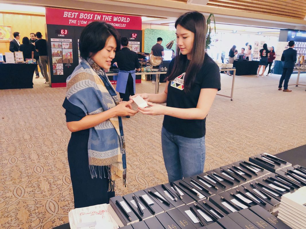 A Partyinkers staff member handing over a custom-engraved prize to a participant at an event, representing the 'Minute to Win It with Custom-Engraved Trophies' section. The table is lined with personalized items, showcasing the unique and rewarding experience for winners of quick, engaging games.
