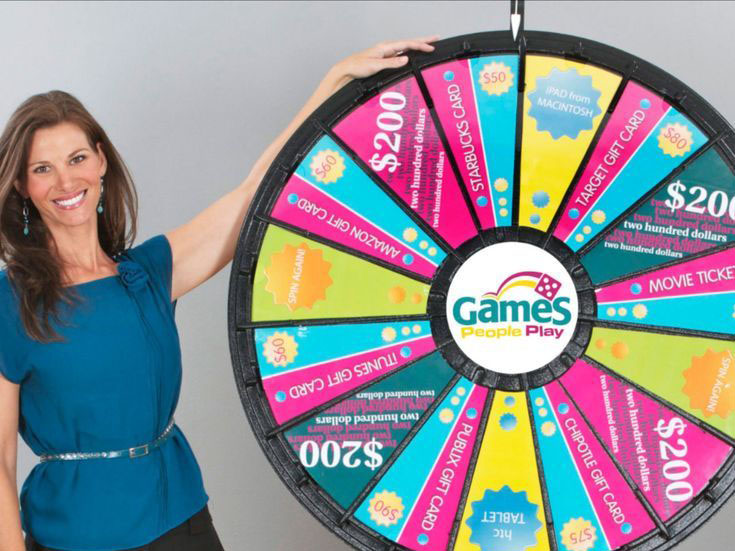 A woman standing next to a brightly colored prize wheel with various rewards, including gift cards and cash prizes. The wheel, labeled 'Games People Play,' represents the 'Spin the Wheel with Customized Rewards' section, highlighting the excitement and variety of prizes that attendees can win at Dinner and Dance events.