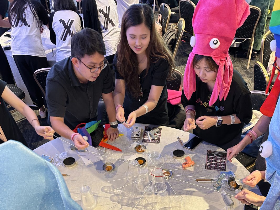 A group of participants working together in a team-building activity at an event, focusing on a collaborative task involving strings and objects. Some members are wearing fun, themed hats, adding a playful element to the challenge. This image represents the 'Team-Building Activities with Onsite Customization' section, where teamwork and personalization combine to create memorable experiences.