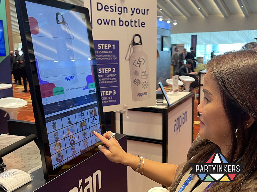 An attendee at a conference using a touchscreen kiosk to design a personalized aluminum bottle with custom graphics, provided by Partyinkers' live digital printing service.