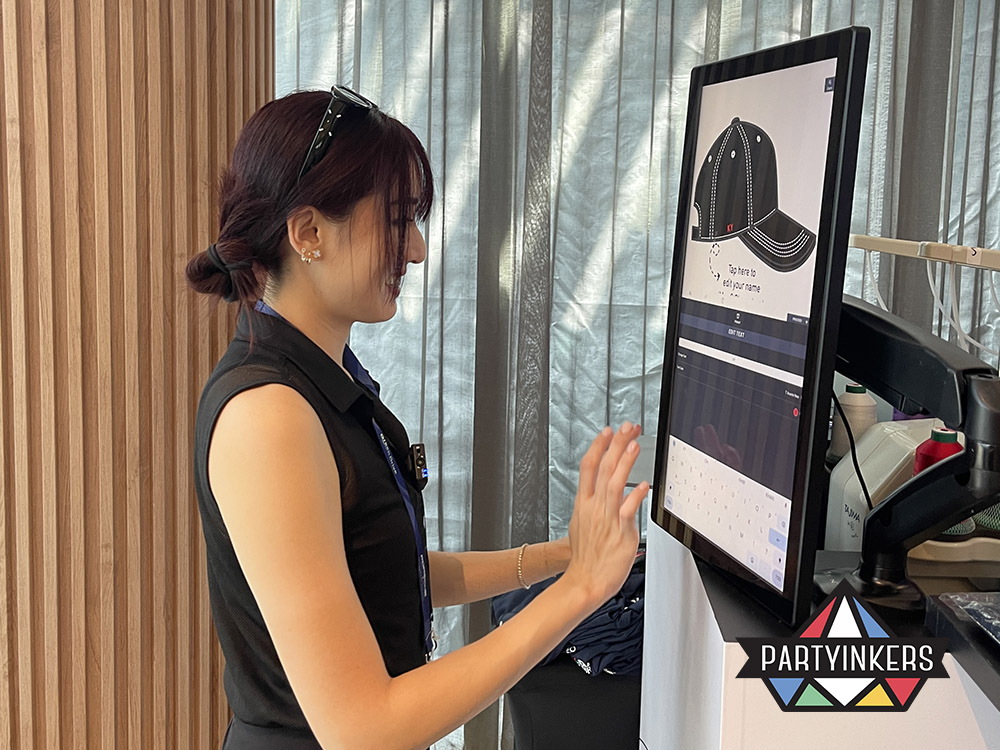 An event attendee using a touchscreen kiosk to design and personalize an embroidered cap with custom text, provided by Partyinkers' live embroidery service.