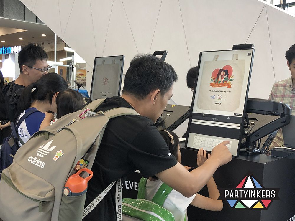 Attendees at a live printing booth designing personalized tote bags at a corporate family day event, with Partyinkers offering interactive on-site customization.