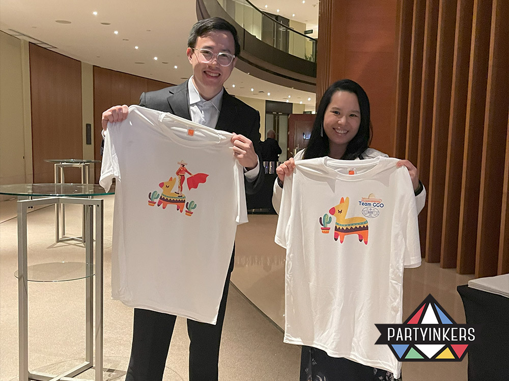 Two event attendees proudly holding up custom-designed t-shirts they received as branded merchandise at a corporate family day activities, showcasing the personalized gifts offered by Partyinkers.
