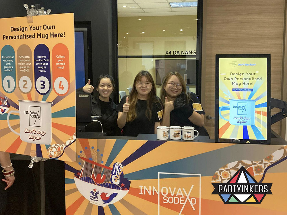 Three event staff members smiling and giving thumbs up at a live personalized mug printing station, showcasing the customization process at a corporate family day event. The booth displays a step-by-step guide for designing and collecting personalized mugs.