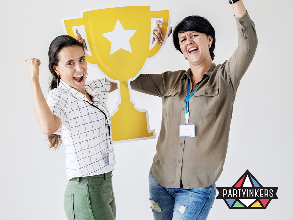 Two women celebrating and holding a large trophy cutout at a corporate family day recognition awards ceremony, showcasing their excitement and team spirit at the event organized by Partyinkers.