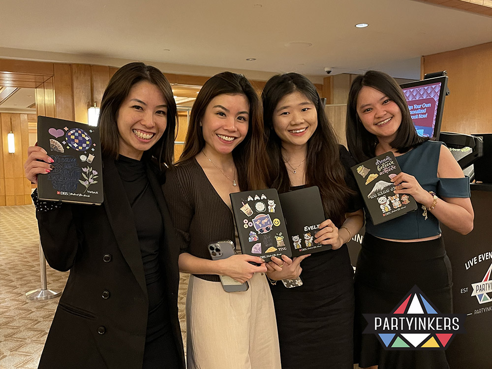 Four smiling attendees proudly display their personalized notebooks created at the Partyinkers interactive booth during an event. The customized designs highlight how interactive stations foster event engagement and provide memorable keepsakes, enhancing the overall attendee experience.