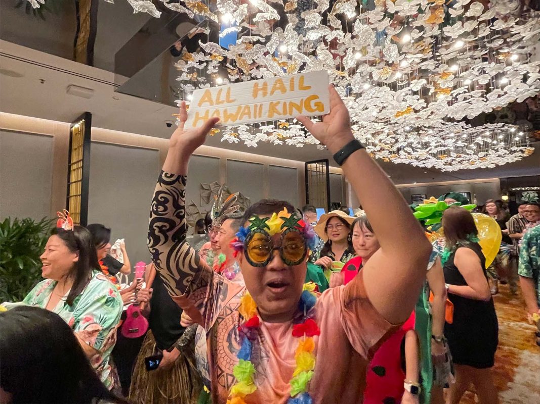A lively attendee at a themed event holding a custom sign that reads 'All Hail Hawaii King,' surrounded by others in festive costumes, showcasing the excitement and engagement that customized elements at interactive booth ideas bring to events