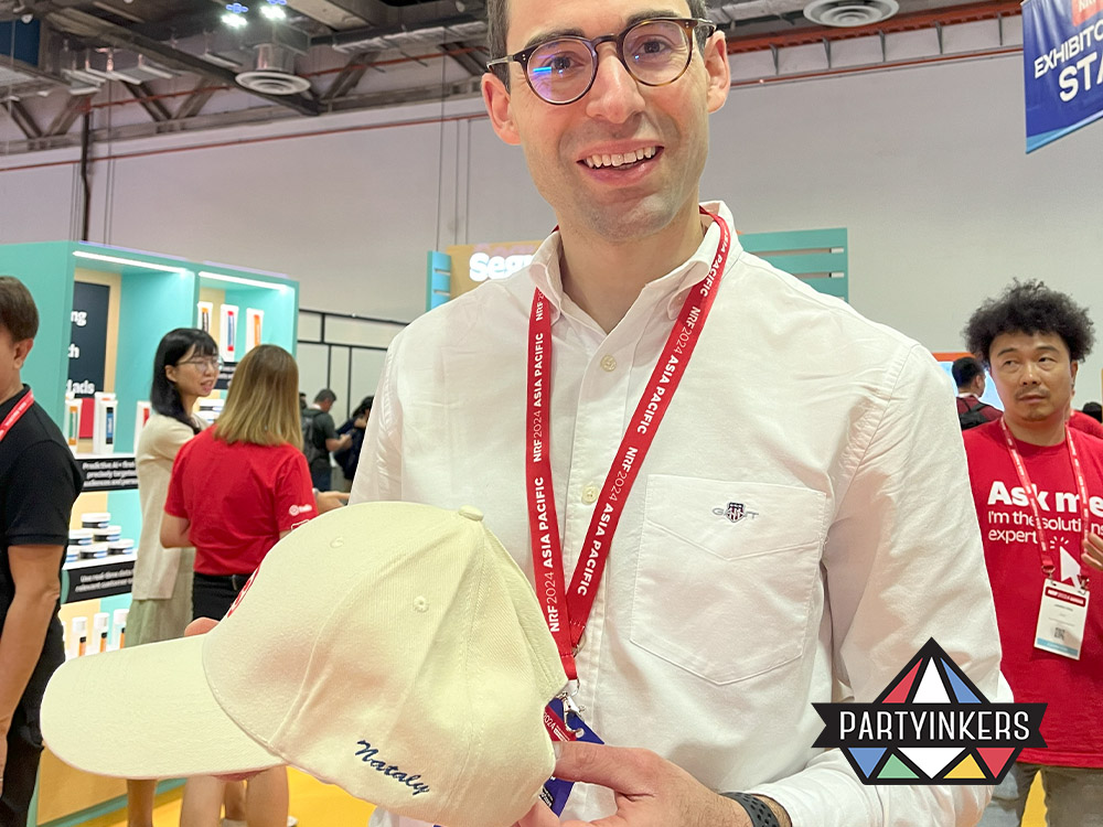 A satisfied attendee at a twilio trade show holding a custom-embroidered cap with his name on it, demonstrating the wide appeal and personalized touch of live embroidery across various events.

