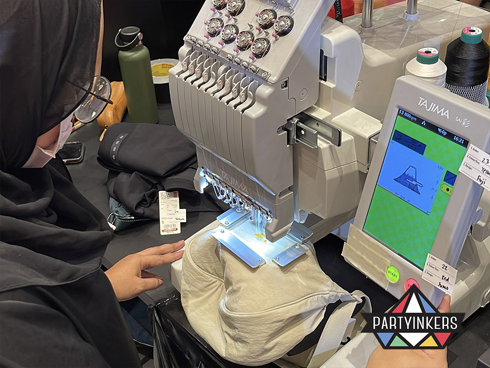 A Partyinkers staff member operating a live embroidery machine to stitch a custom design onto a tote bag, showcasing the intricate craftsmanship and personalized experience provided by live embroidery as an interactive booth ideas at an event.
