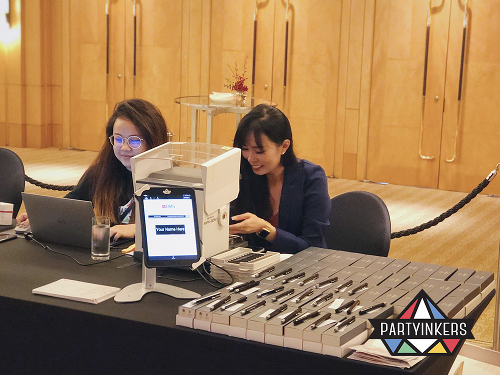 Partyinkers staff preparing custom laser-engraved pens at an event, with a display of completed pens ready for attendees, showcasing the precision and appeal of laser engraving as a standout interactive booth feature.