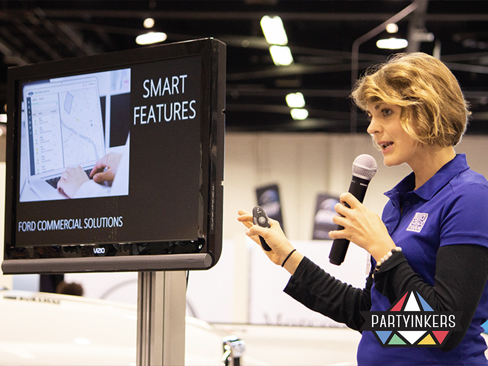 A female presenter delivering a live product demonstration at a trade show booth, showcasing smart features of Ford Commercial Solutions on a screen. The speaker is using a microphone and holding a clicker, engaging the audience with an interactive presentation. The booth setup includes a large display screen showing the product's interface and features, emphasizing the importance of live demos in attracting attendees and demonstrating complex products effectively.