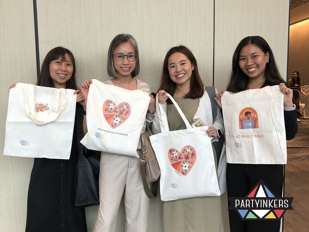 Four smiling women proudly hold up their personalized tote bags featuring unique and colorful designs, showcasing one of the "Trade Show Booth Ideas" of providing custom SWAG. The tote bags highlight the value of live printing services at trade show booths, allowing attendees to create memorable and useful branded items, enhancing their overall event experience.
