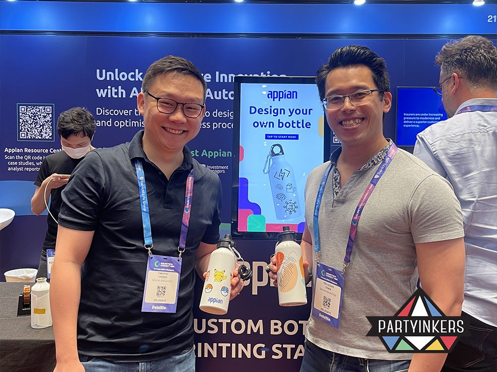Two event attendees hold customized water bottles designed at a live printing station during a trade show. The station, featuring a digital display with "Design your own bottle" instructions, highlights the interactive nature of Partyinkers' booth setup, offering visitors a memorable hands-on experience.
