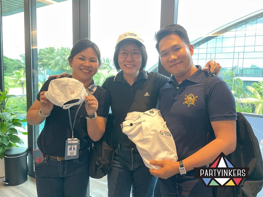 Three smiling individuals proudly display personalized apparel, including caps and polo shirts, at a trade show event. This image highlights one of the "Trade Show Booth Ideas" of offering custom-branded SWAG. The items showcase the effectiveness of live printing services, creating memorable and high-quality giveaways that attendees can use and cherish, reinforcing brand recognition.