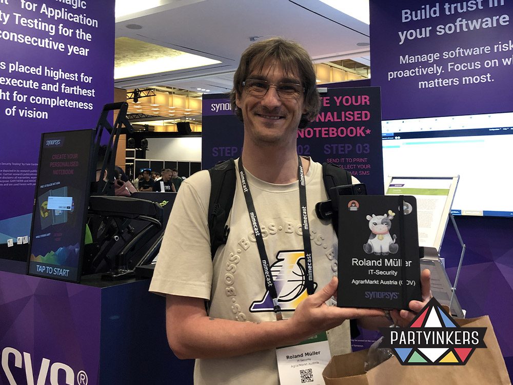 A smiling man proudly holds a personalized notebook at a trade show booth, illustrating one of the "Trade Show Booth Ideas" of offering custom-branded items. The booth's vibrant setup in the background highlights the interactive element of live printing services, allowing attendees to create unique, memorable souvenirs that align with their professional interests and event experience.