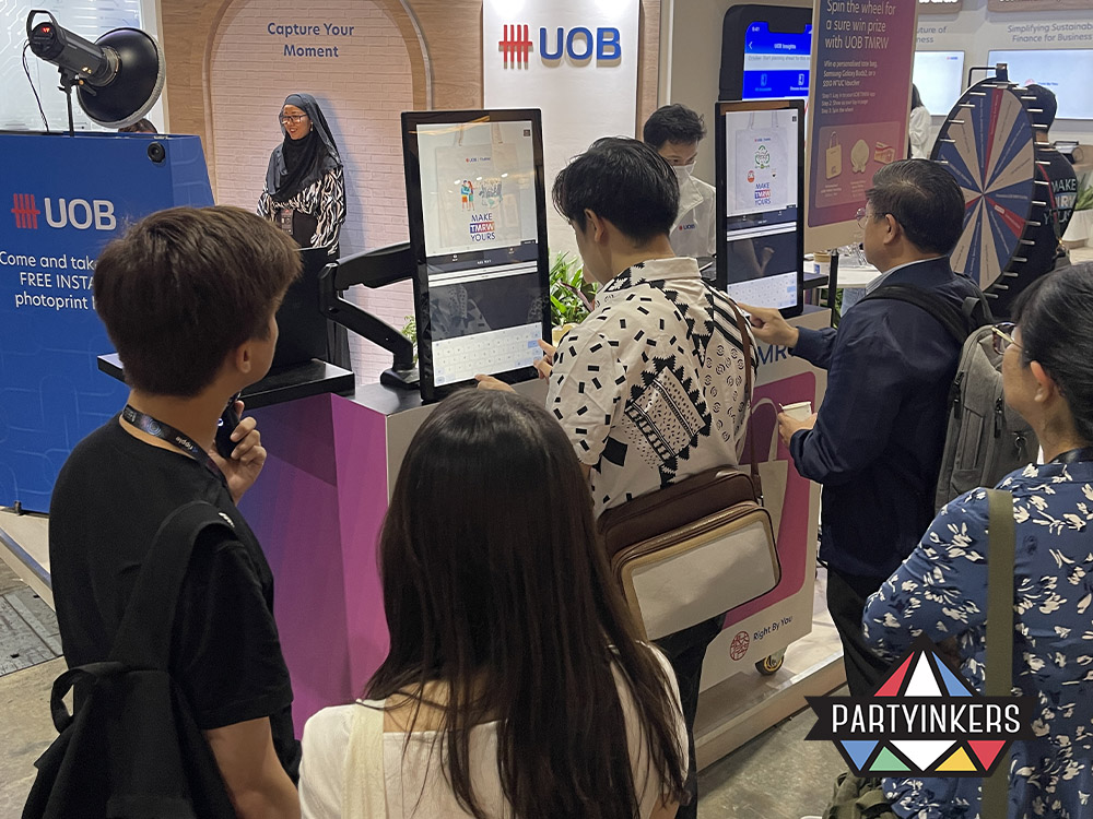 Attendees interacting with live digital printing kiosks at the UOB booth during an event, showcasing interactive event ideas such as personalized tote bag printing. The booth attracted participants through hands-on activities, enhancing engagement and brand experience.
