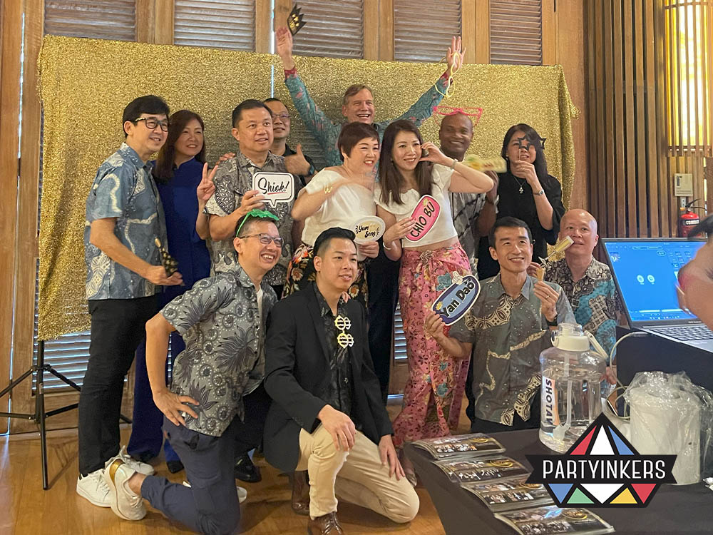 "A cheerful group of attendees posing with fun photo props against a gold backdrop at a Partyinkers-hosted event, emphasizing a lively and engaging atmosphere."