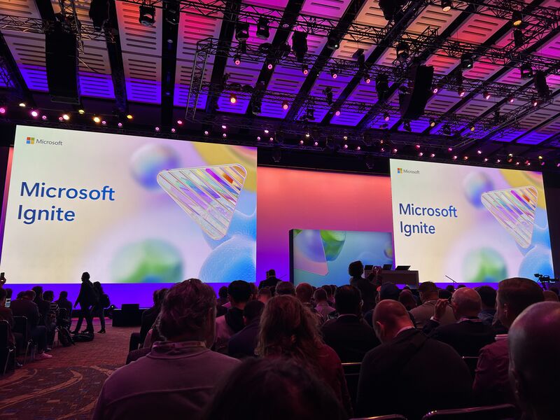 Attendees at the Microsoft Ignite conference are captivated by a vibrant display featuring the event's branding on large screens. The setting is a spacious conference hall filled with professionals from around the globe, gathered to explore the latest innovations and insights in technology offered by Microsoft