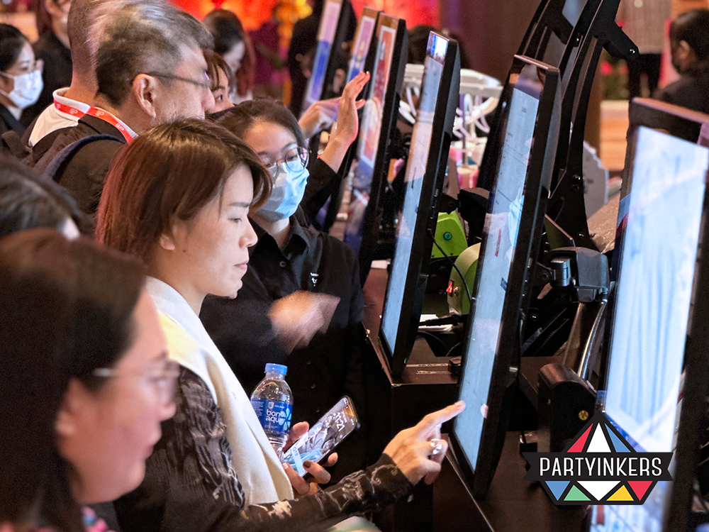 Attendees engage actively at a customization station, using touchscreen panels to select designs for personalizing t-shirts and other merchandise. The station is busy with participants and staff, creating a dynamic atmosphere at the event.