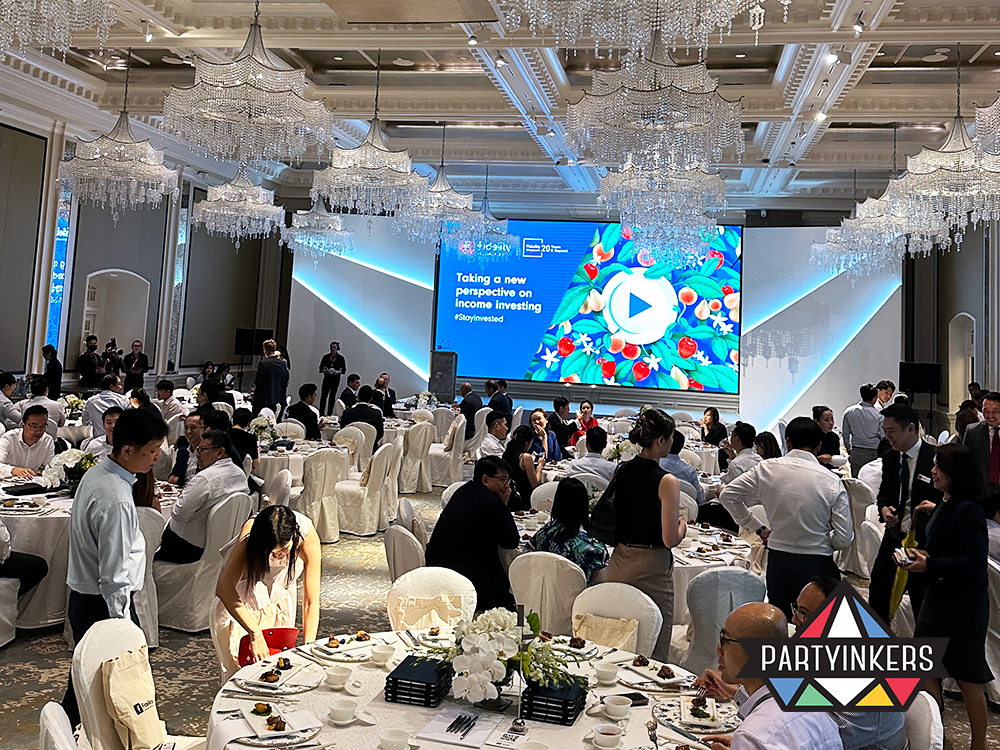 Professionals network at a high-profile conference focused on new perspectives on income investing, hosted in a grand ballroom adorned with crystal chandeliers. The event features round tables set for dining, a large presentation screen displaying the conference theme, and attendees engaged in discussions, highlighting the blend of formal presentation and interactive opportunities.