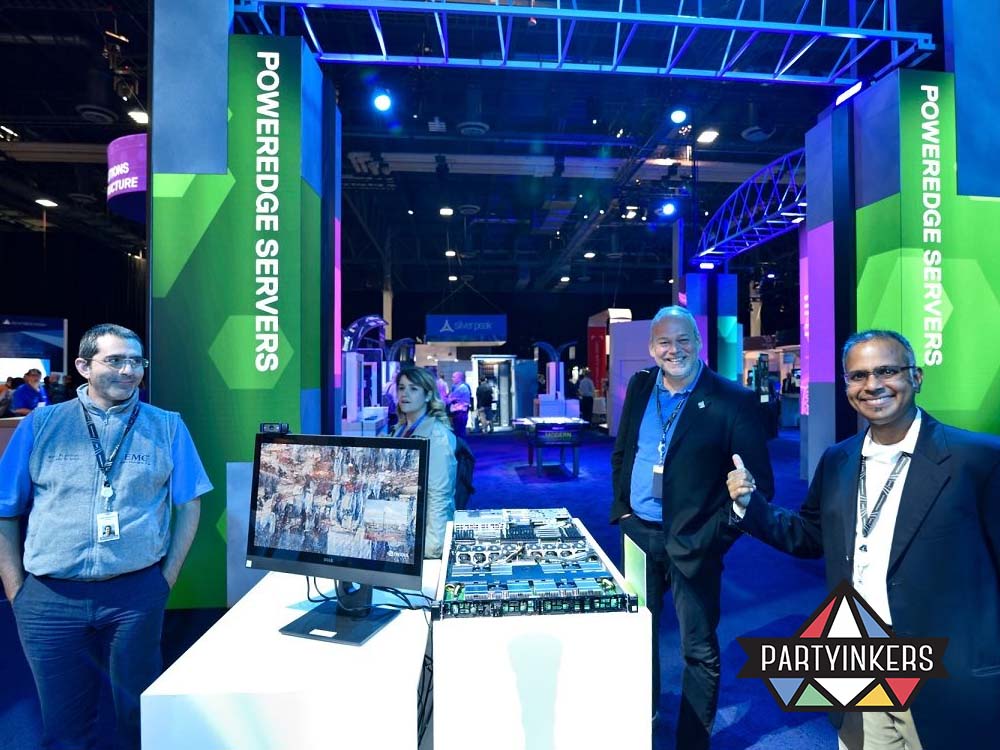 Three professionals standing at an exhibit booth showcasing Dell Technology, with a monitor and server hardware displayed on a table.