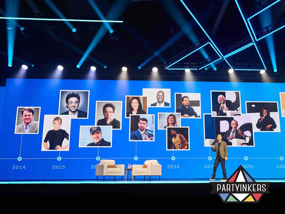 At the X4 Experience Management Summit, a dynamic conference stage with a large screen displaying a timeline of influential speakers and notable figures, illuminated by vibrant blue and white lighting. A presenter stands on the stage near two chairs, adding a professional and engaging atmosphere.