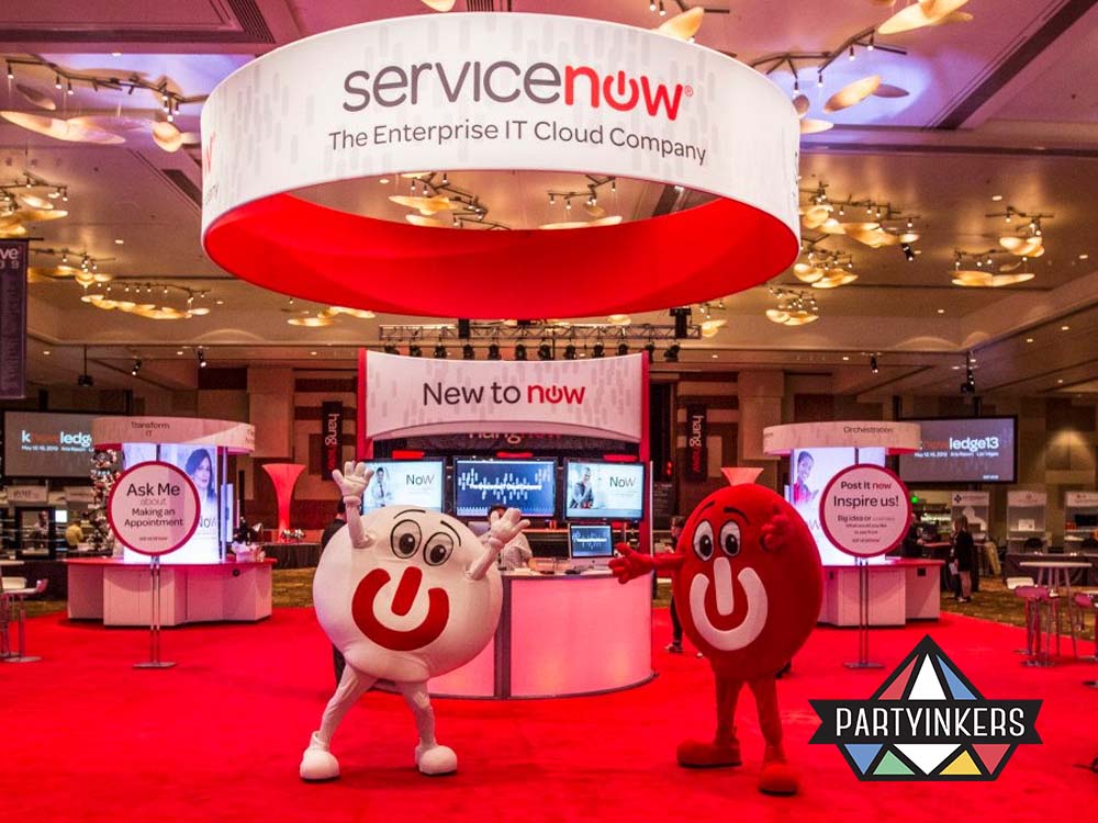 The ServiceNow event booth showcasing a vibrant red and white theme with large overhead branding and interactive stations. Mascots in ServiceNow-themed costumes engage attendees in the lively exhibition space.