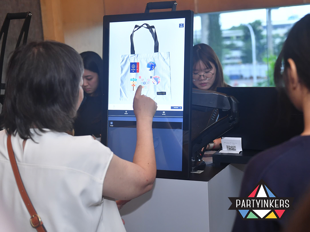A participant at the American Express event uses the touchscreen interface to design a custom tote bag featuring various travel-themed clipart. Partyinkers staff assists in the background, ensuring a seamless personalization experience.