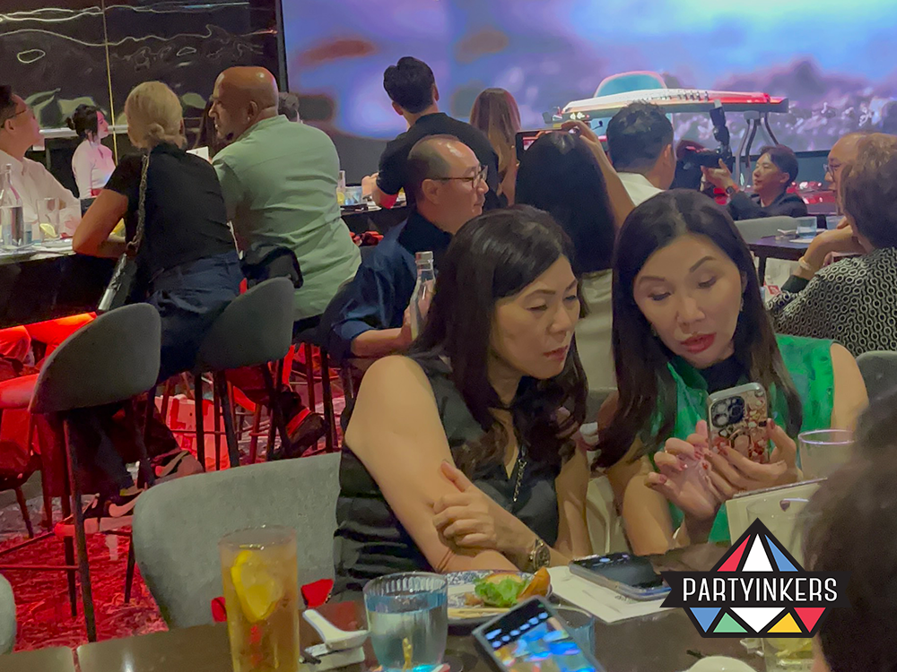 Two elegantly dressed women engage deeply with a smartphone at a VIP networking event, surrounded by a diverse group of attendees in a sophisticated dining setting. They appear to be sharing insights or coordinating efforts, highlighting the active collaboration and high-level engagement typical at exclusive social gatherings.