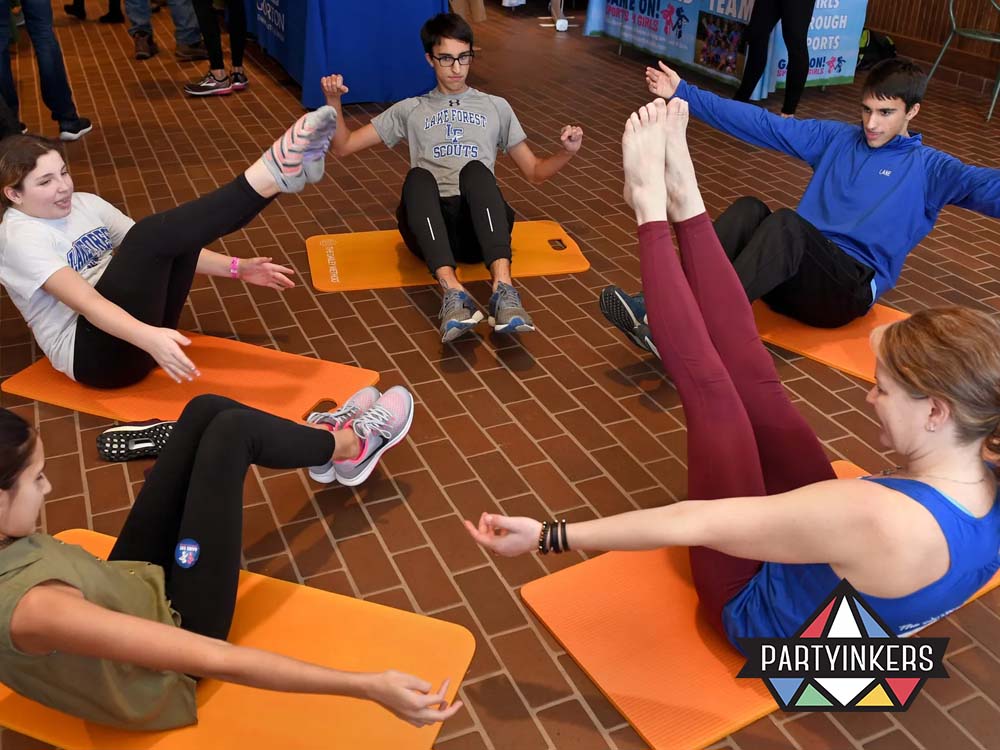 A group of individuals participating in a pop-up fitness session, performing seated leg raise exercises on orange mats in a communal indoor space.