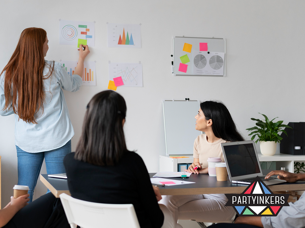 A professional development workshop in progress, featuring a diverse group of participants focused on a presenter. The presenter, a young woman with red hair, points to various charts on a wall, explaining data analysis techniques. The setting is a well-organized office space, conducive to interactive learning and discussion