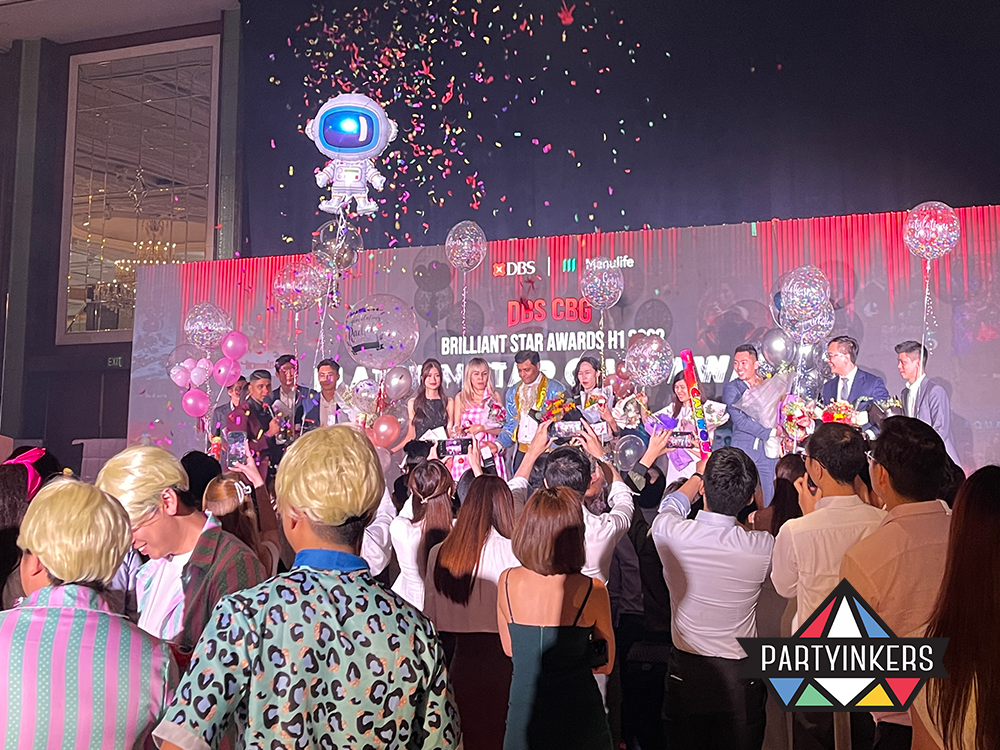 "An awards ceremony with a confetti-filled celebration, featuring a stage packed with awardees, balloons, and a festive crowd applauding their achievements."