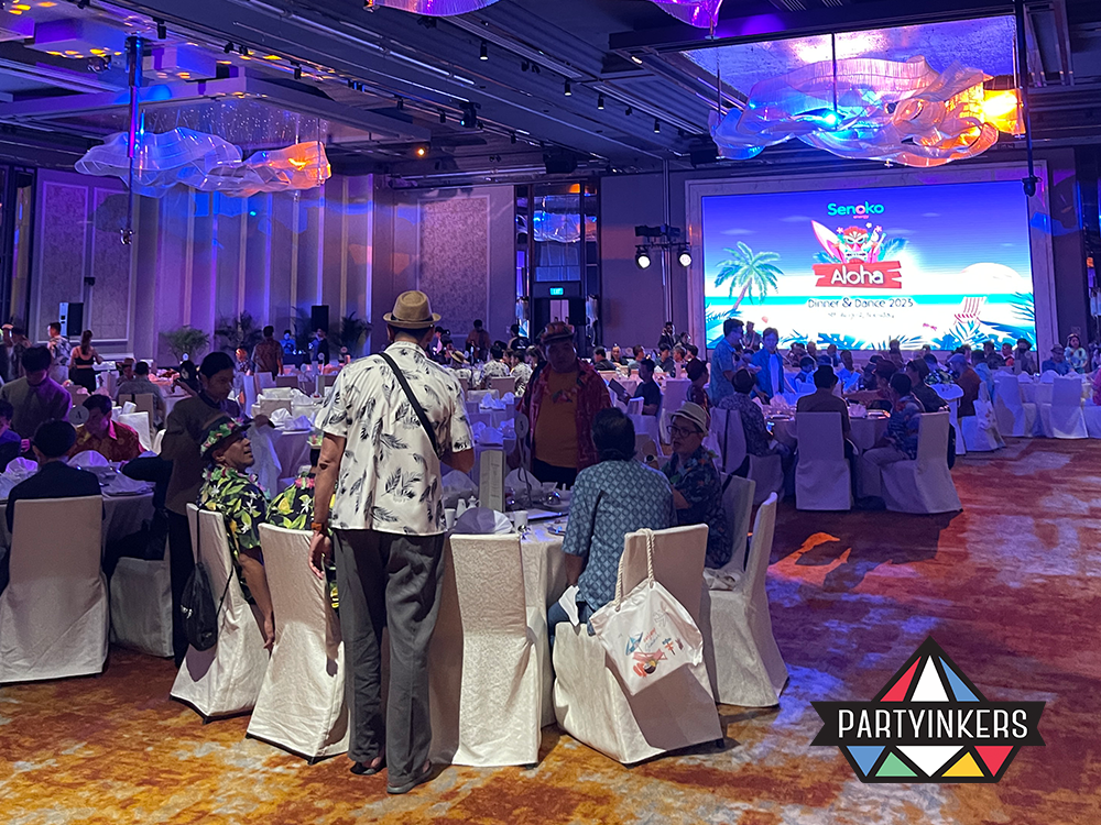 "A lively dinner and dance event with an 'Aloha' theme, featuring vibrant decorations, round tables, and engaged participants dressed in tropical attire."