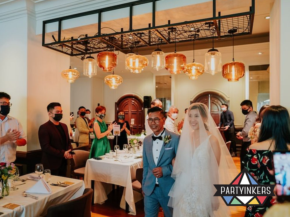 A joyful wedding celebration inside an elegant dining room, with the newlyweds walking in amidst warm applause from guests seated at beautifully decorated tables.