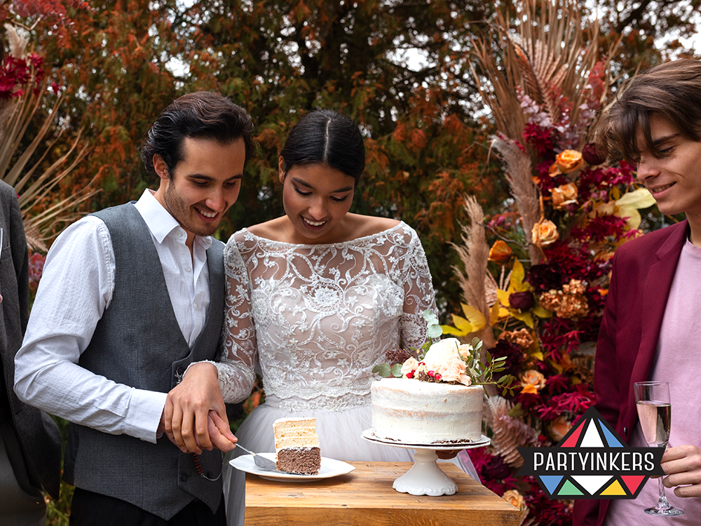 "A newlywed couple cutting a rustic wedding cake outdoors, surrounded by autumn-themed floral arrangements and friends sharing in the moment."