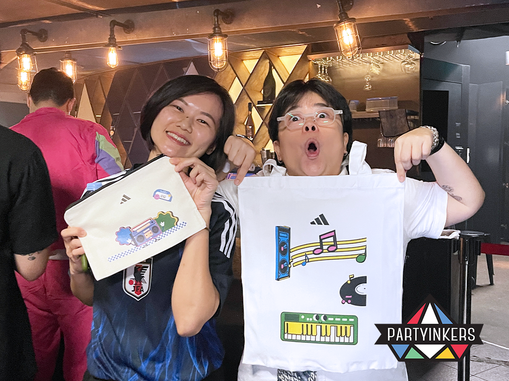 "Two event attendees showcasing personalized tote bags and pouches with vibrant, creative designs printed live at a Partyinkers booth, highlighting the event's interactive and artistic focus."