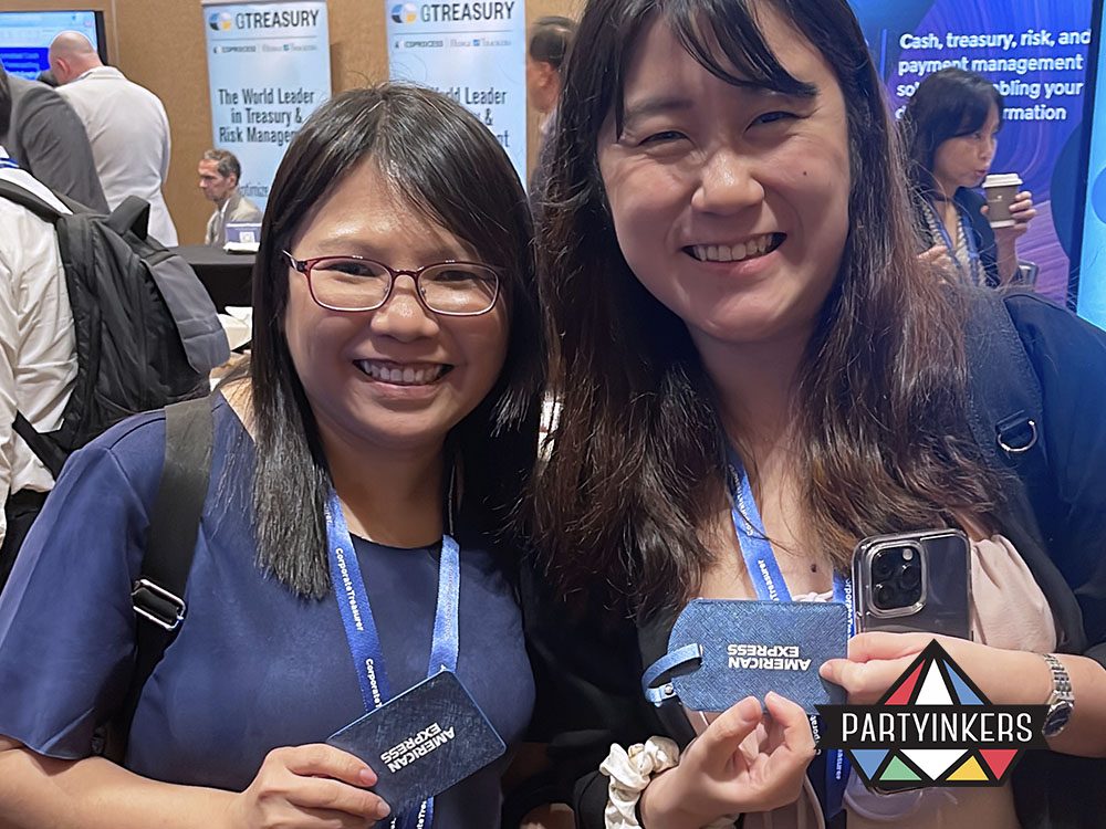 Two smiling attendees showcasing their personalized luggage tags branded with "American Express" at a professional event.