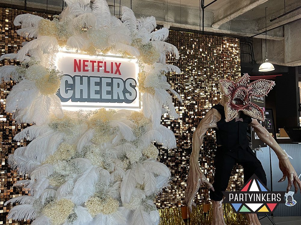 A dramatic display at the Netflix Cheers event featuring a Demogorgon prop and a white feathered backdrop with glittering gold accents.