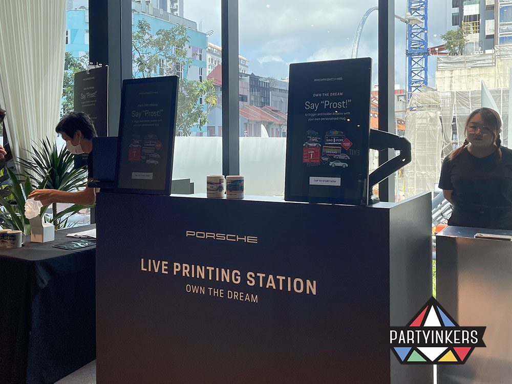 A sleek Porsche live printing station set up at an event, featuring interactive screens and equipment for creating personalized merchandise.