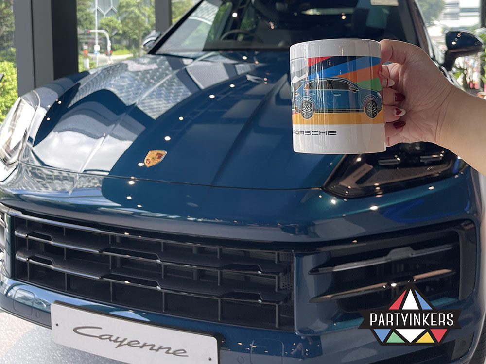 A custom Porsche-themed mug held in front of a shiny blue Porsche Cayenne, highlighting personalized automotive branding.