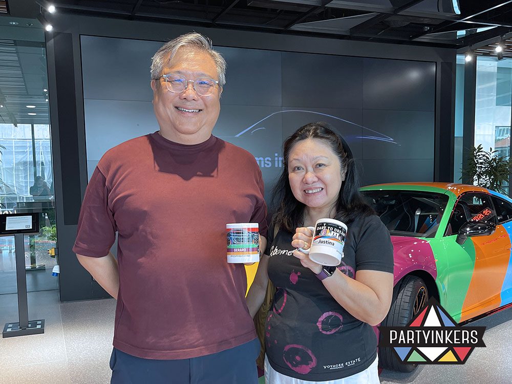 A happy couple showcasing their customized Porsche-themed mugs at a vibrant event venue with a colorful car in the background.